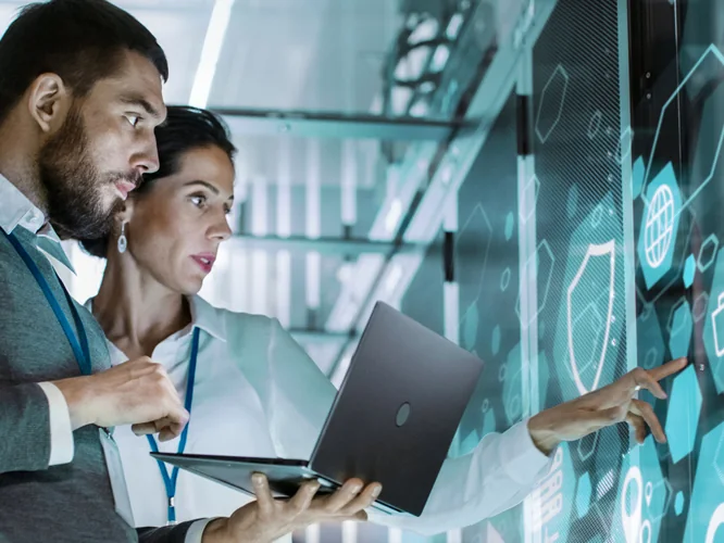 Consultores de TI com notebook analisando um painel digital de segurança e nuvem no data center